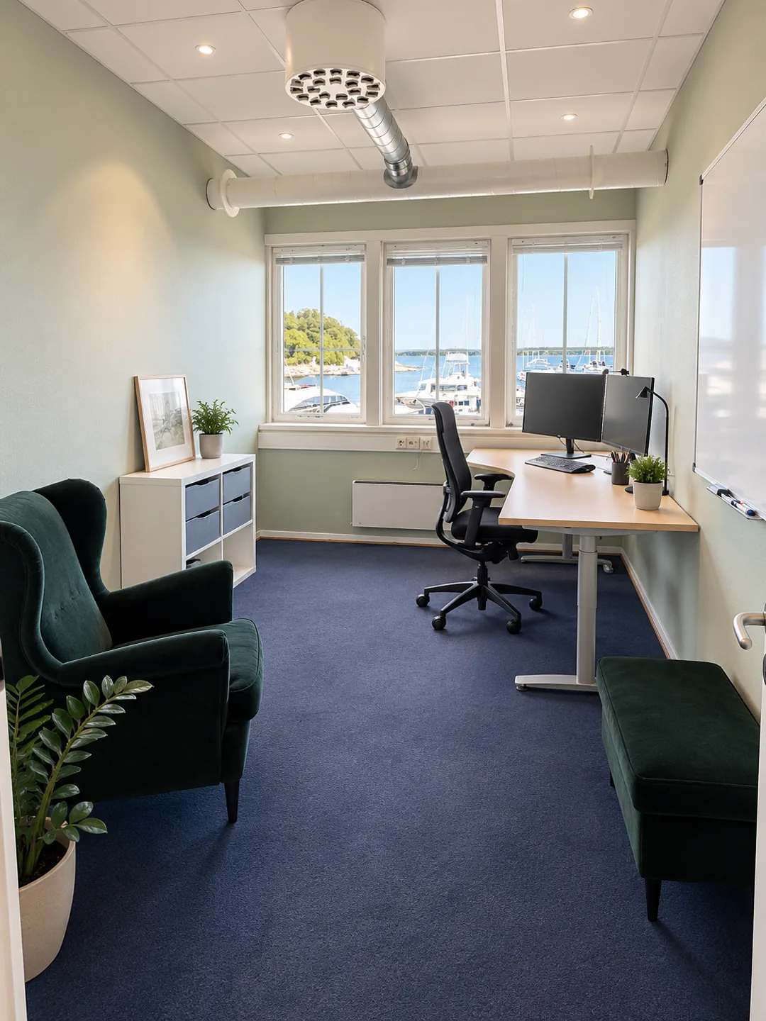 Møblert cellekontor med arbeidsbord, skjermer og grønne lenestoler ved vinduene